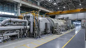 Vista interior de una instalación de generación de energía con equipos industriales, tuberías metálicas y una grúa puente.